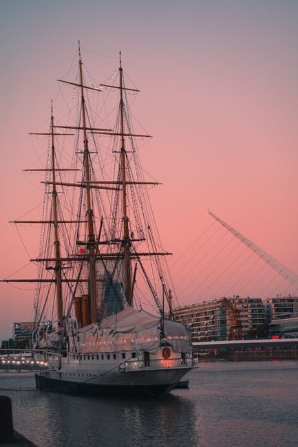 Majestic sailing ship docked near a modern bridge at sunrise with a clear sky.