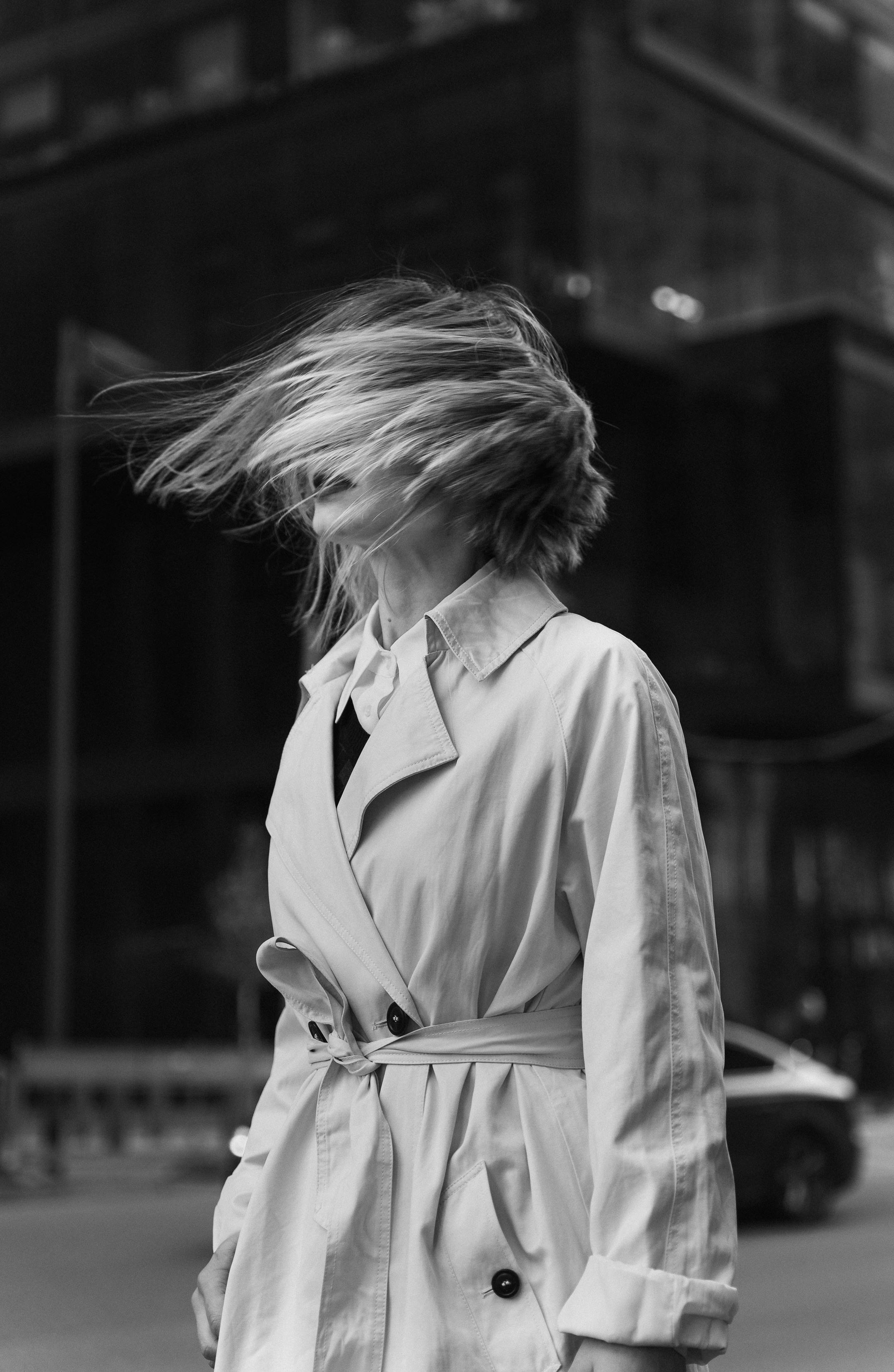 A woman in a trench coat stands in the city as the wind blows her hair dramatically.