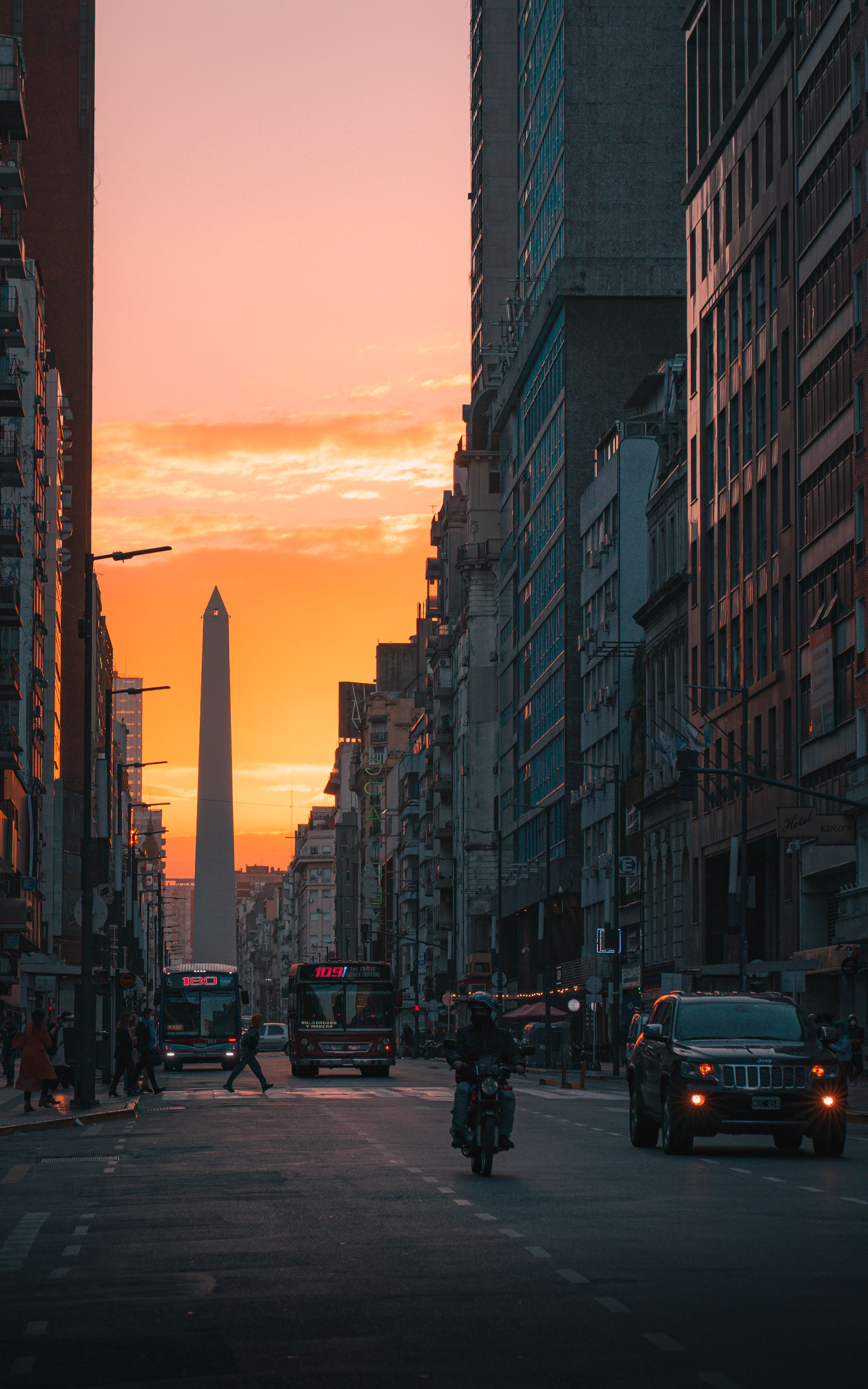 A bustling city street at sunset with a prominent obelisk in the background.