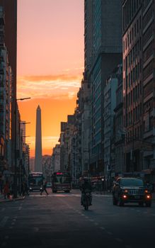 A bustling city street at sunset with a prominent obelisk in the background.