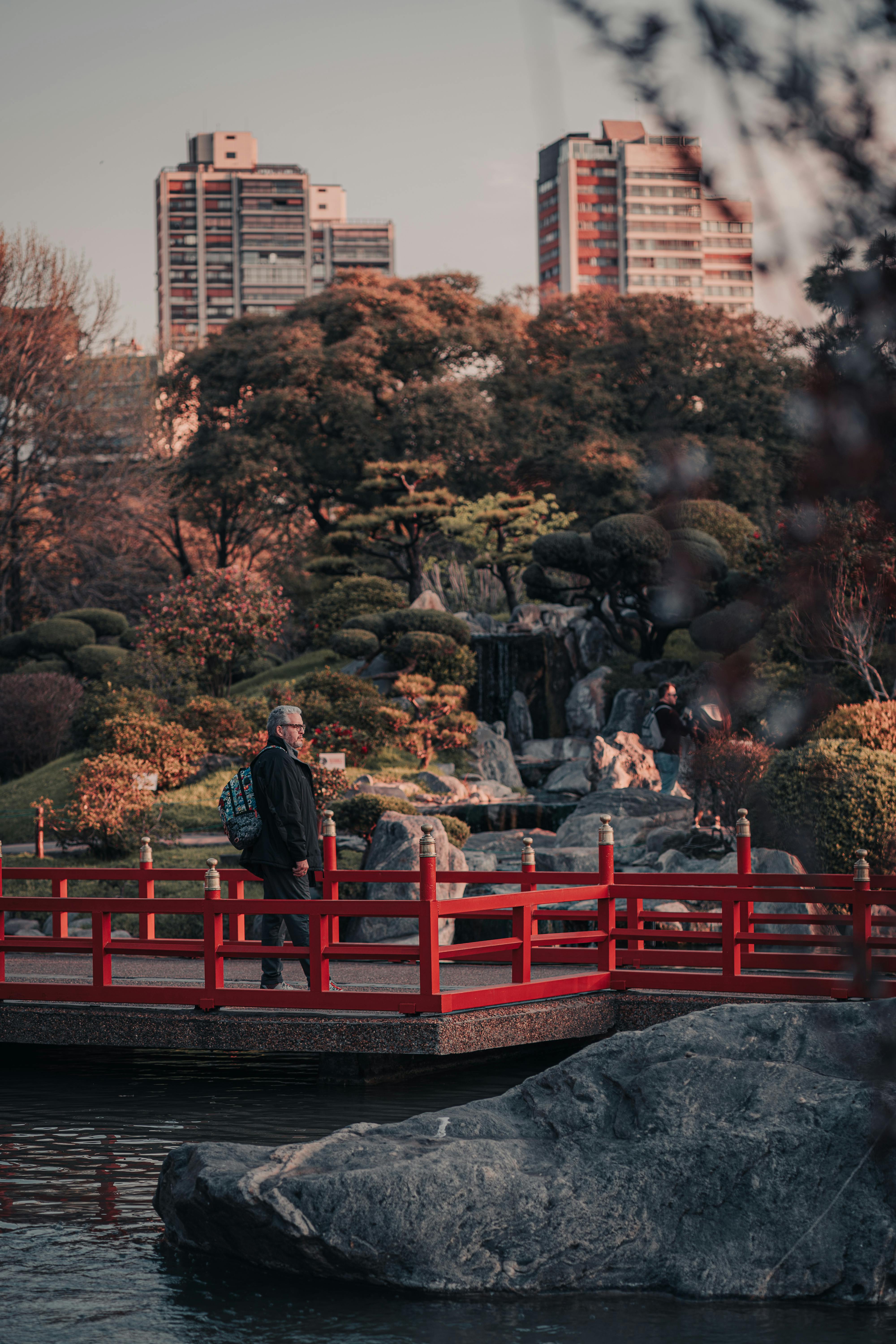 A man strolls across a red bridge in a lush urban park with city buildings in the background.