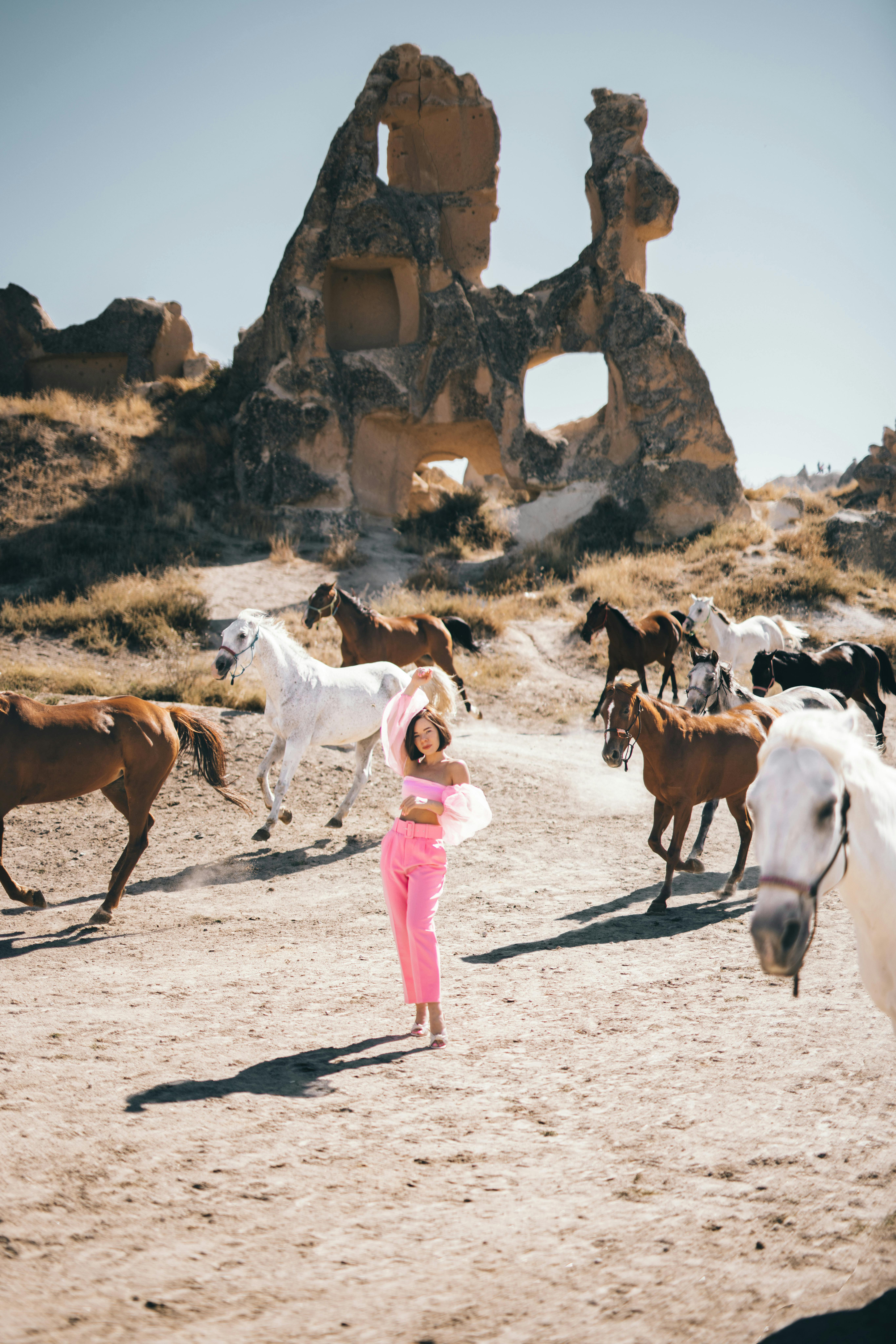 A woman poses among a herd of horses with ancient stone ruins in the backdrop, capturing a vibrant desert scene.