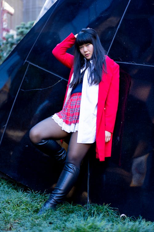 Young Woman in a Plaid Skirt, Boots and Red Jacket Standing against a Black Building 