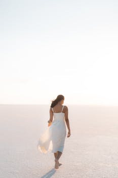 A woman in a flowing white dress walks barefoot across a sunlit salt lake, creating a serene scene.