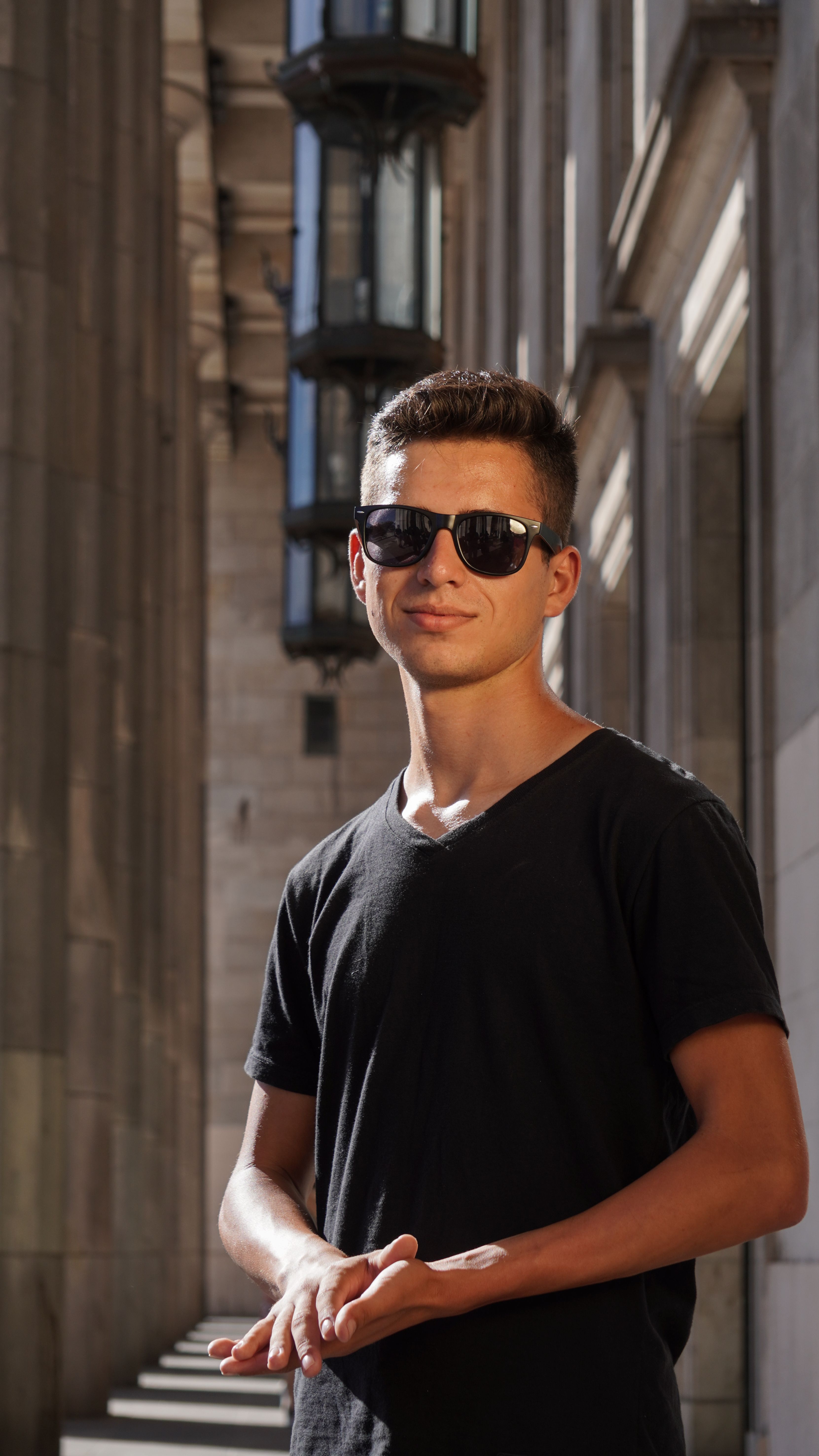 A young man wearing sunglasses smiles confidently in an urban setting, capturing the essence of summer.