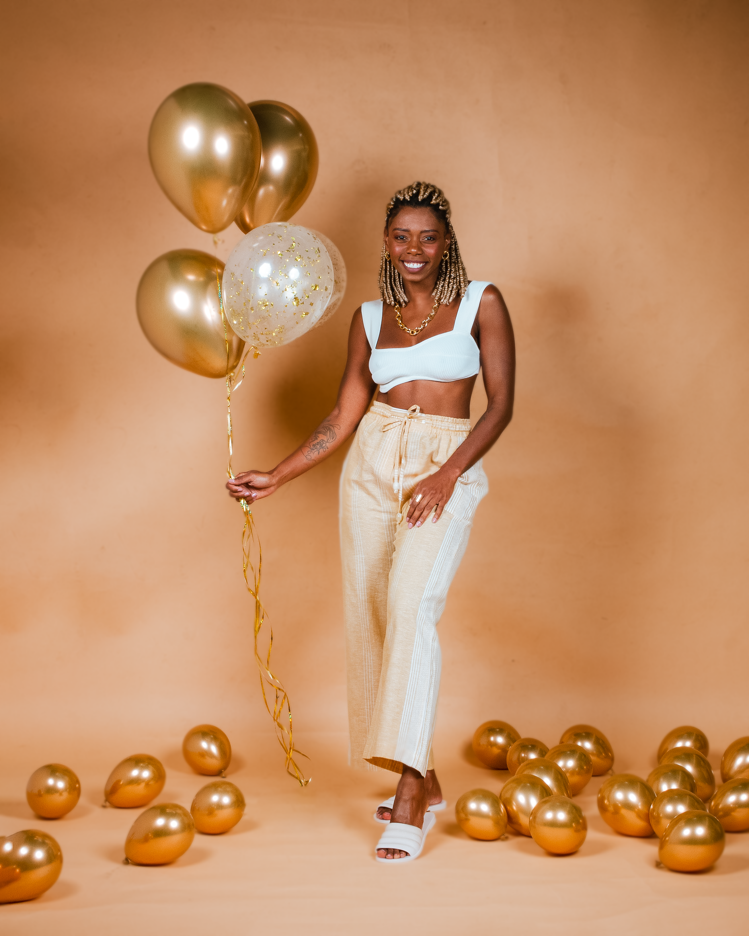 Joyful woman with golden balloons standing indoors against a yellow background, smiling and posing.