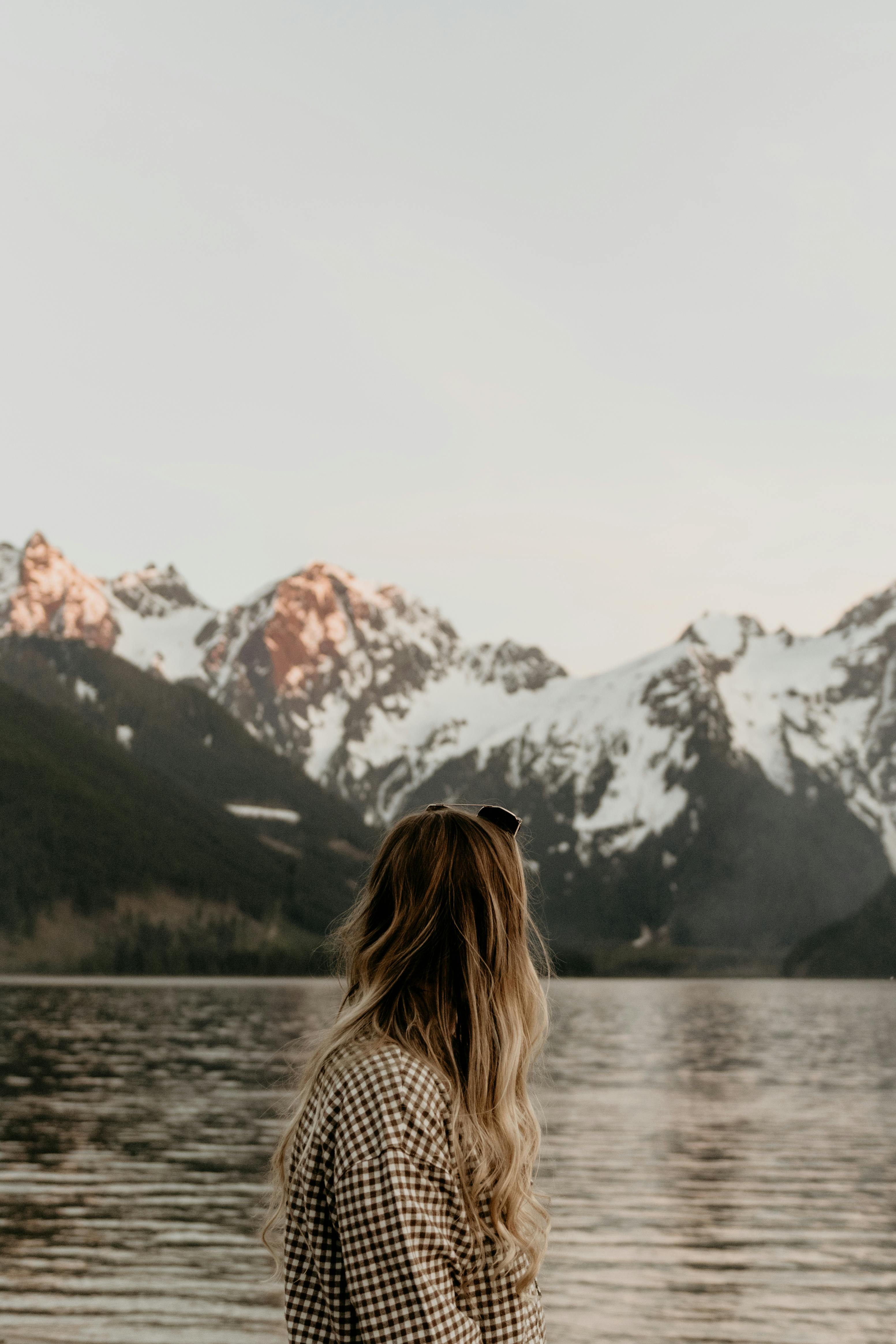 A woman views snowy mountain peaks over a serene lake, surrounded by nature.