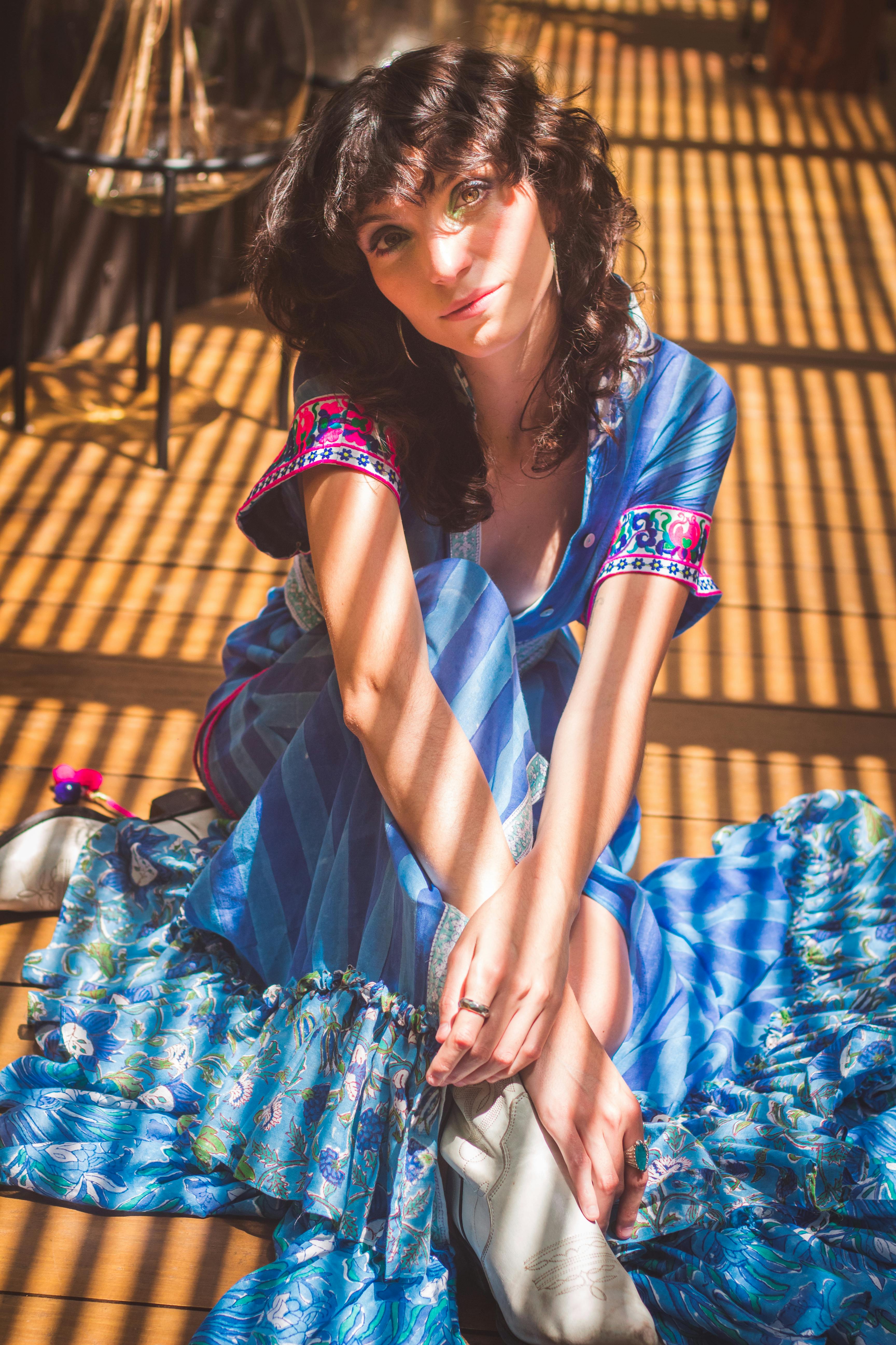 Free A fashionable woman in a blue dress sits with sunlight casting striped shadows indoors. Stock Photo