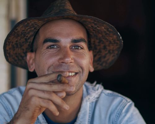 Free A smiling man wearing a straw hat enjoys a cigar in a casual setting, showcasing a relaxed lifestyle. Stock Photo