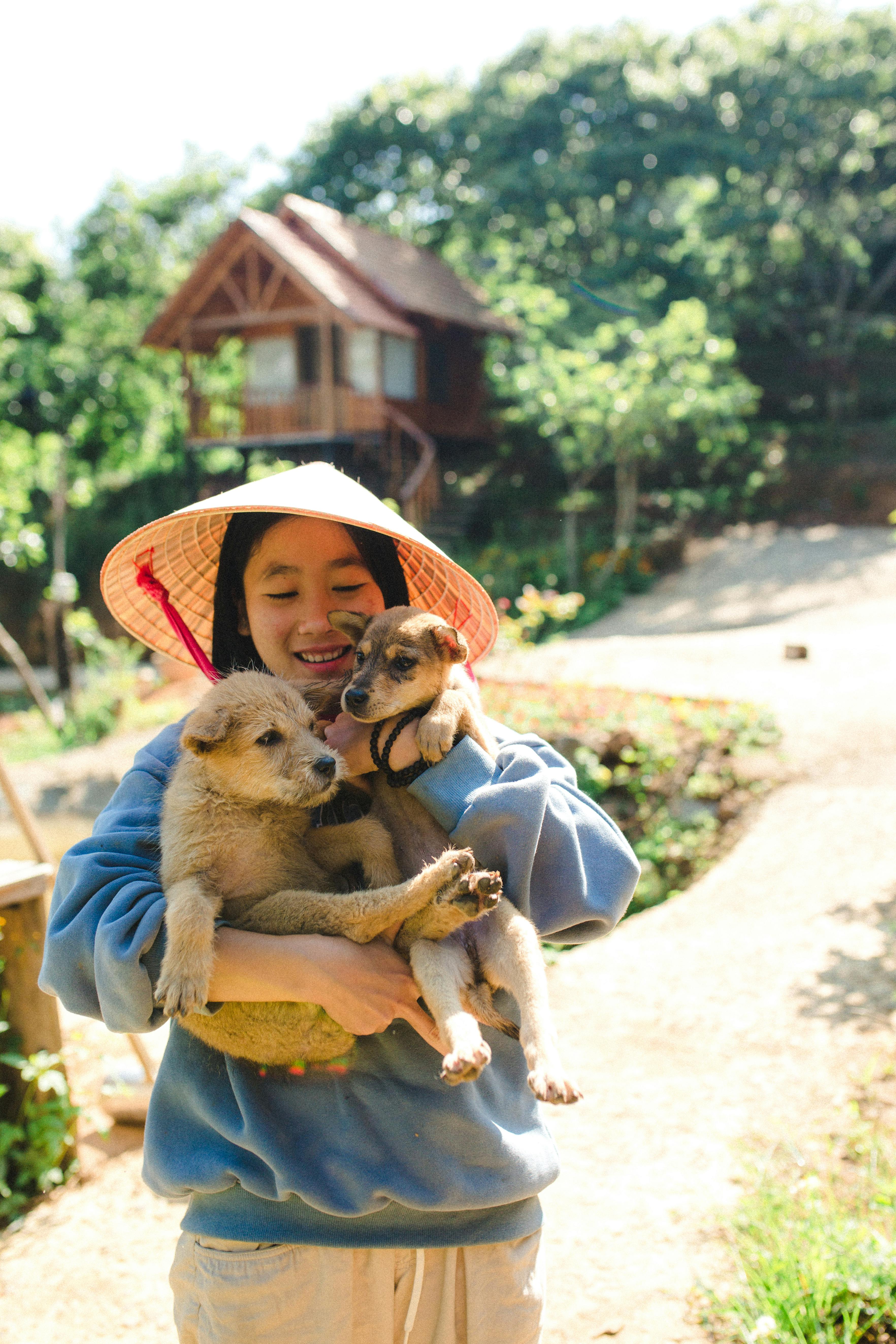 A joyful woman wearing a sunhat holds two cute puppies in a sunny outdoor setting.
