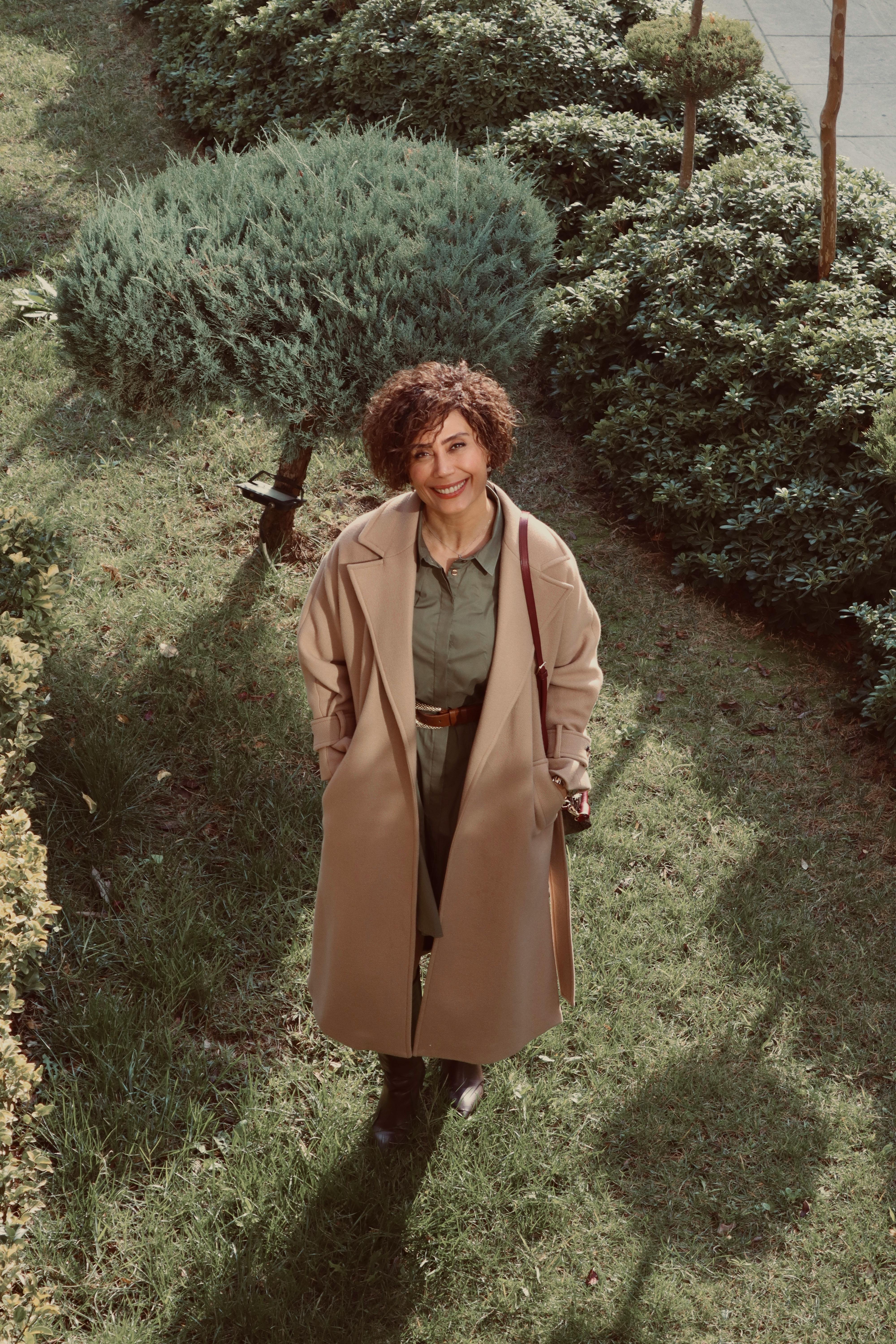 Smiling woman in beige coat standing on green grass in an outdoor garden setting.