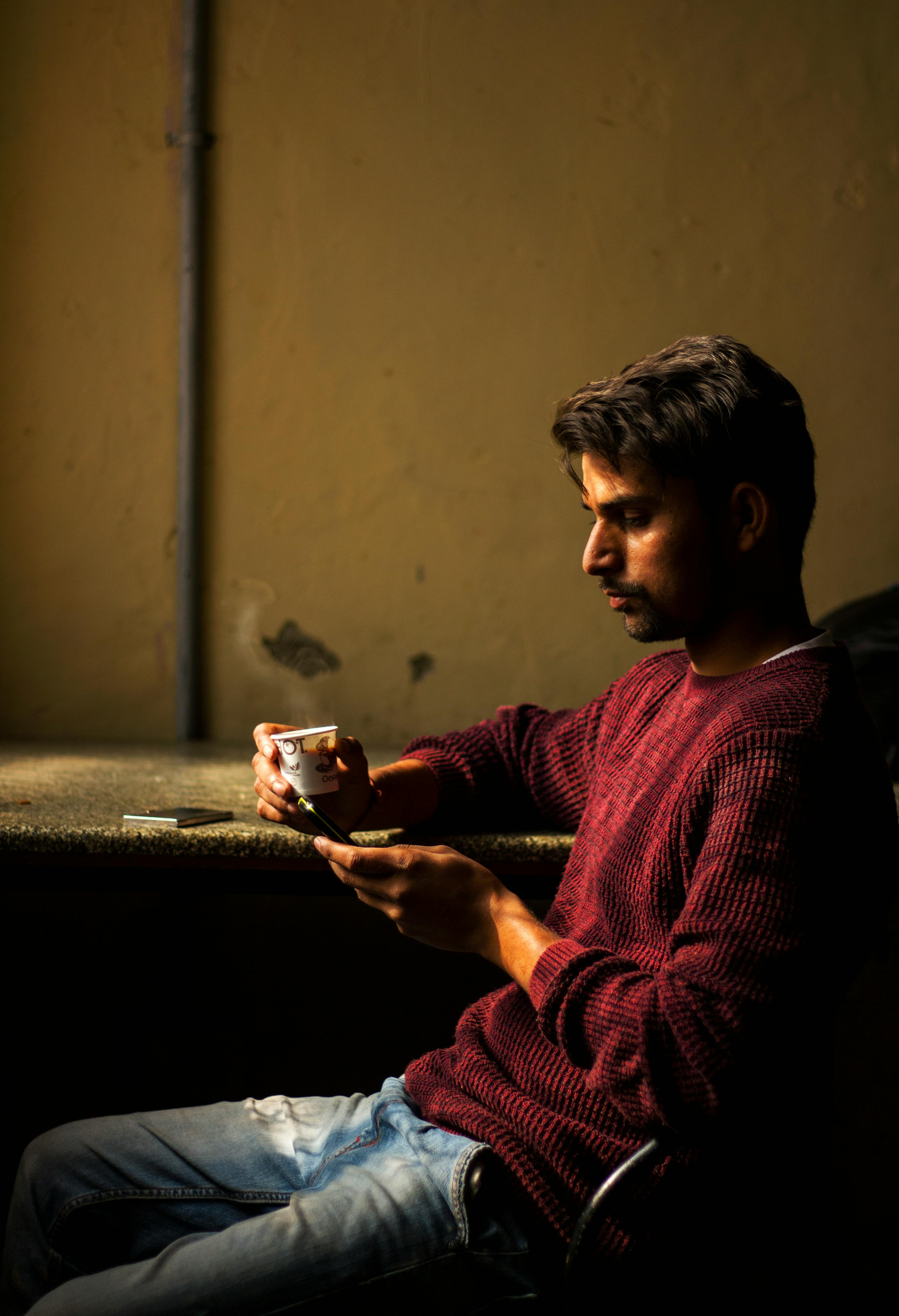 Man Wearing Red Sweater Sitting at Brown Table Holding White Cup