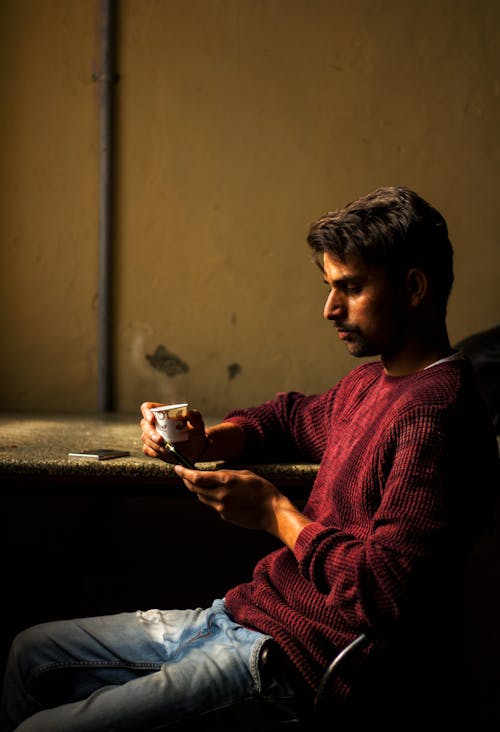 Free Man Wearing Red Sweater Sitting at Brown Table Holding White Cup Stock Photo