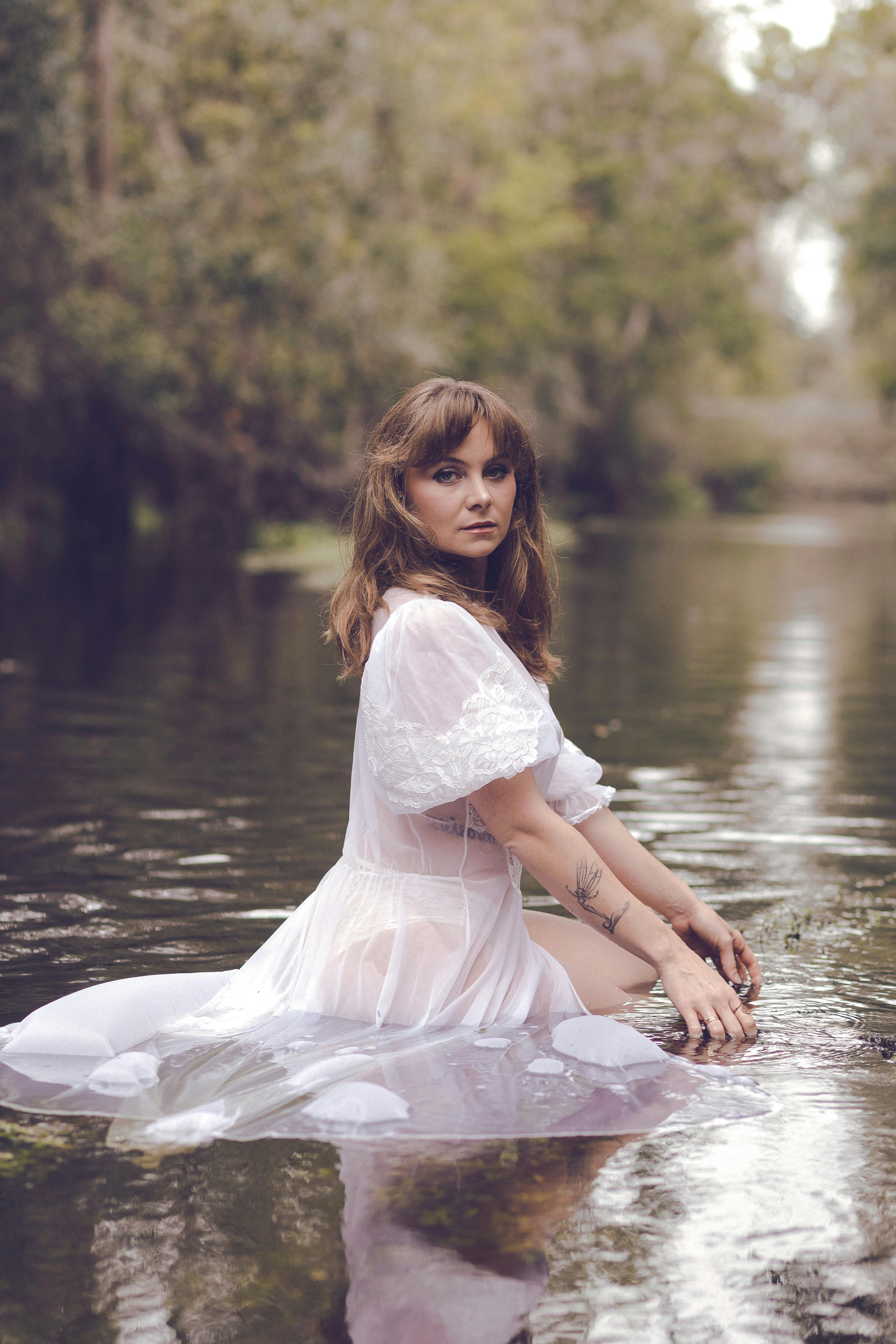 A woman in a delicate white dress poses gracefully in a calm river surrounded by lush forest.