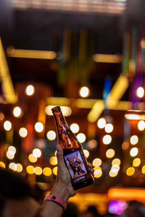 Free A hand holds a beer bottle against a backdrop of colorful bokeh lights indoors at night. Stock Photo