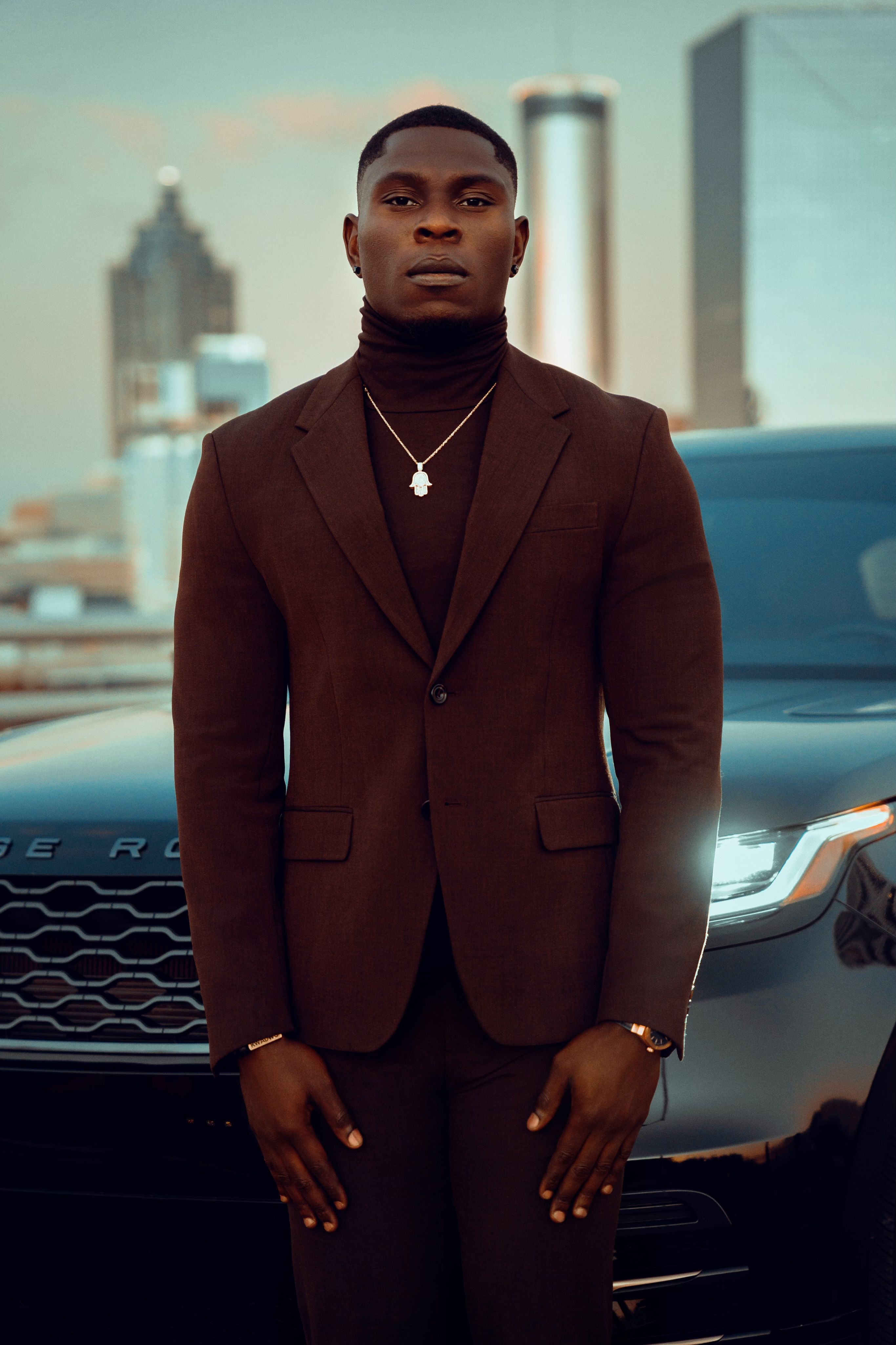 Elegant young man in a brown suit standing in front of a luxury SUV with a city skyline backdrop.