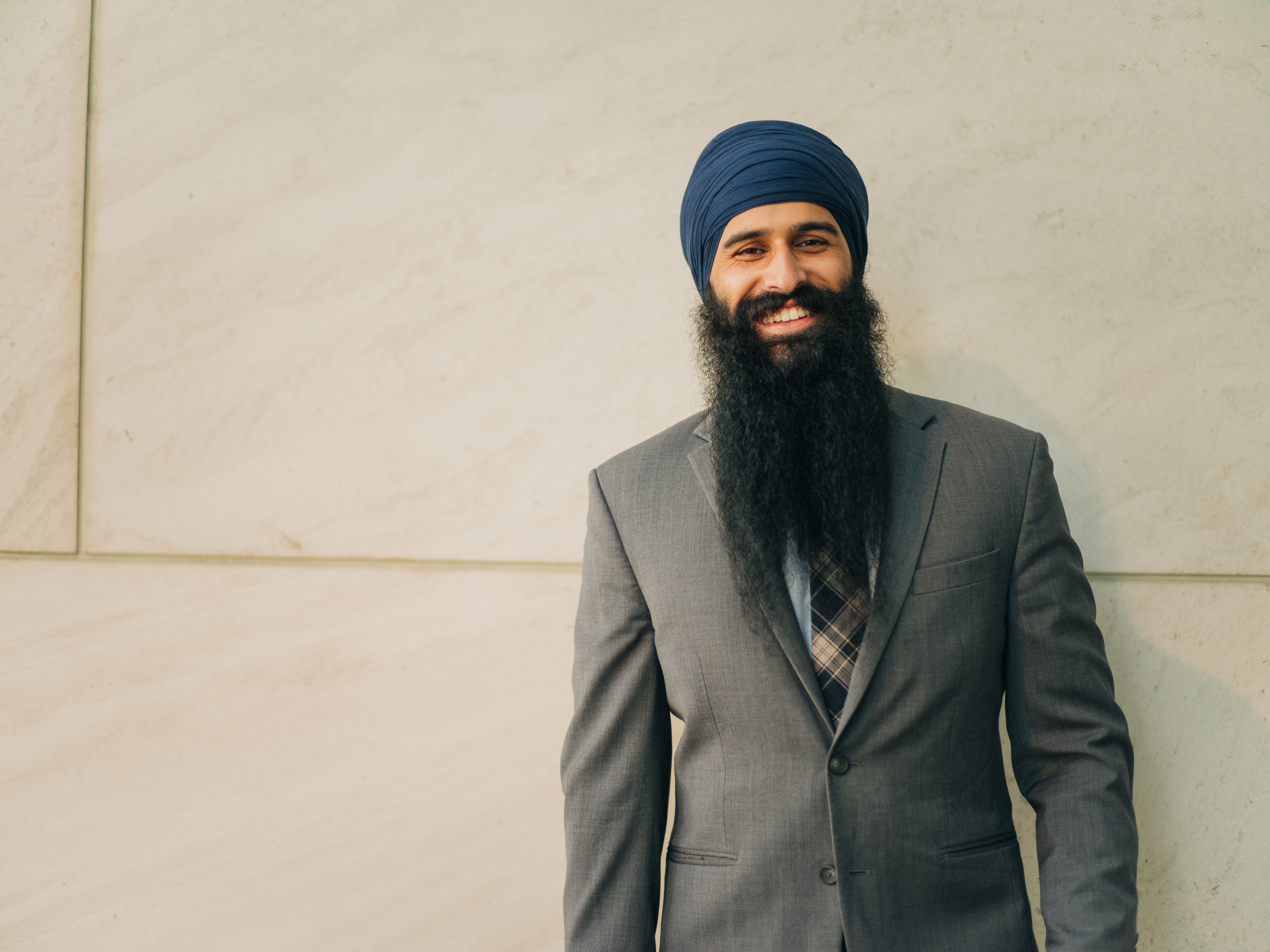 Sikh businessman in full suit and turban smiling confidently against a cream wall.