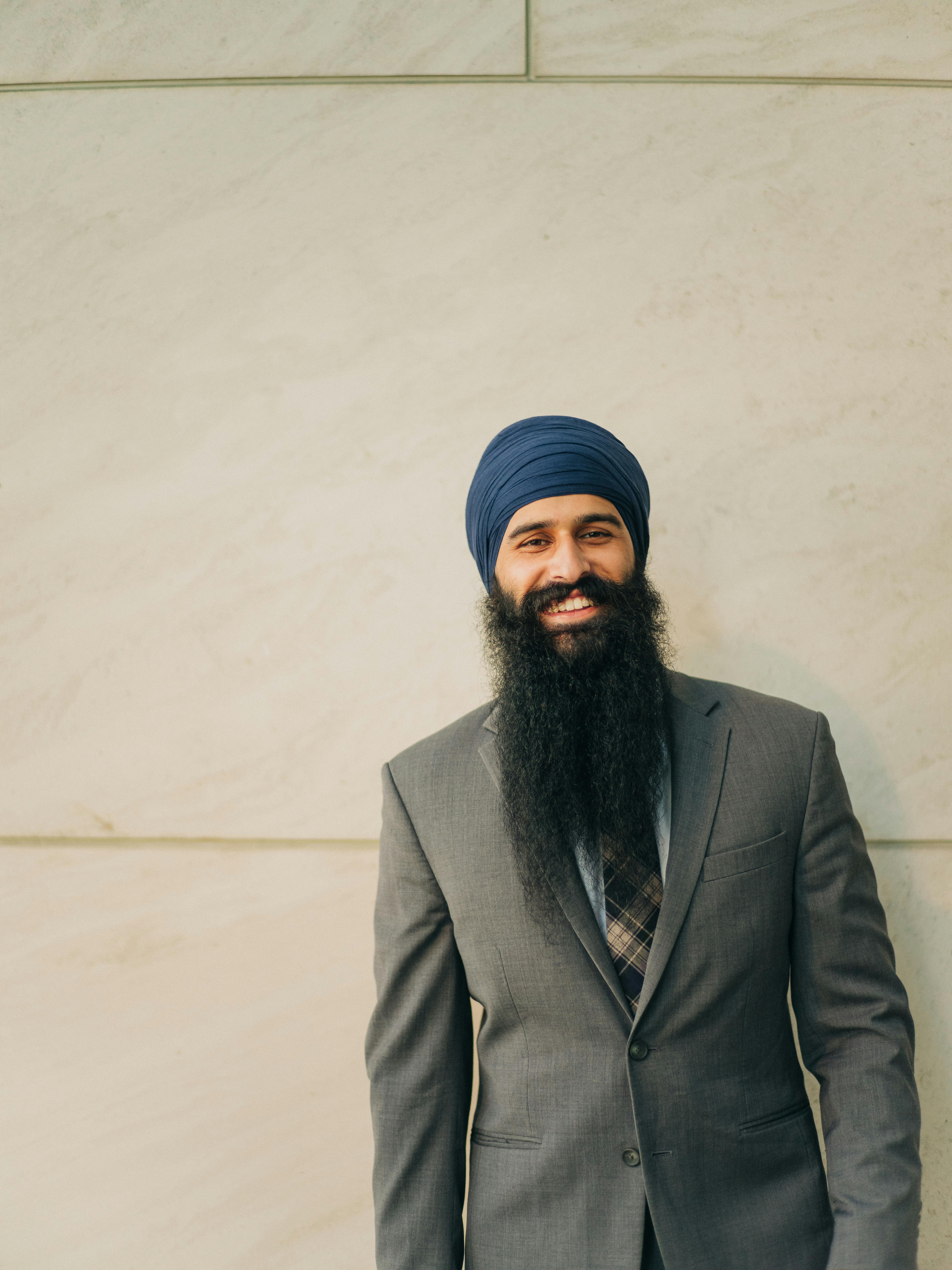 A professional Sikh businessman dressed in a formal suit smiles confidently against a neutral background.