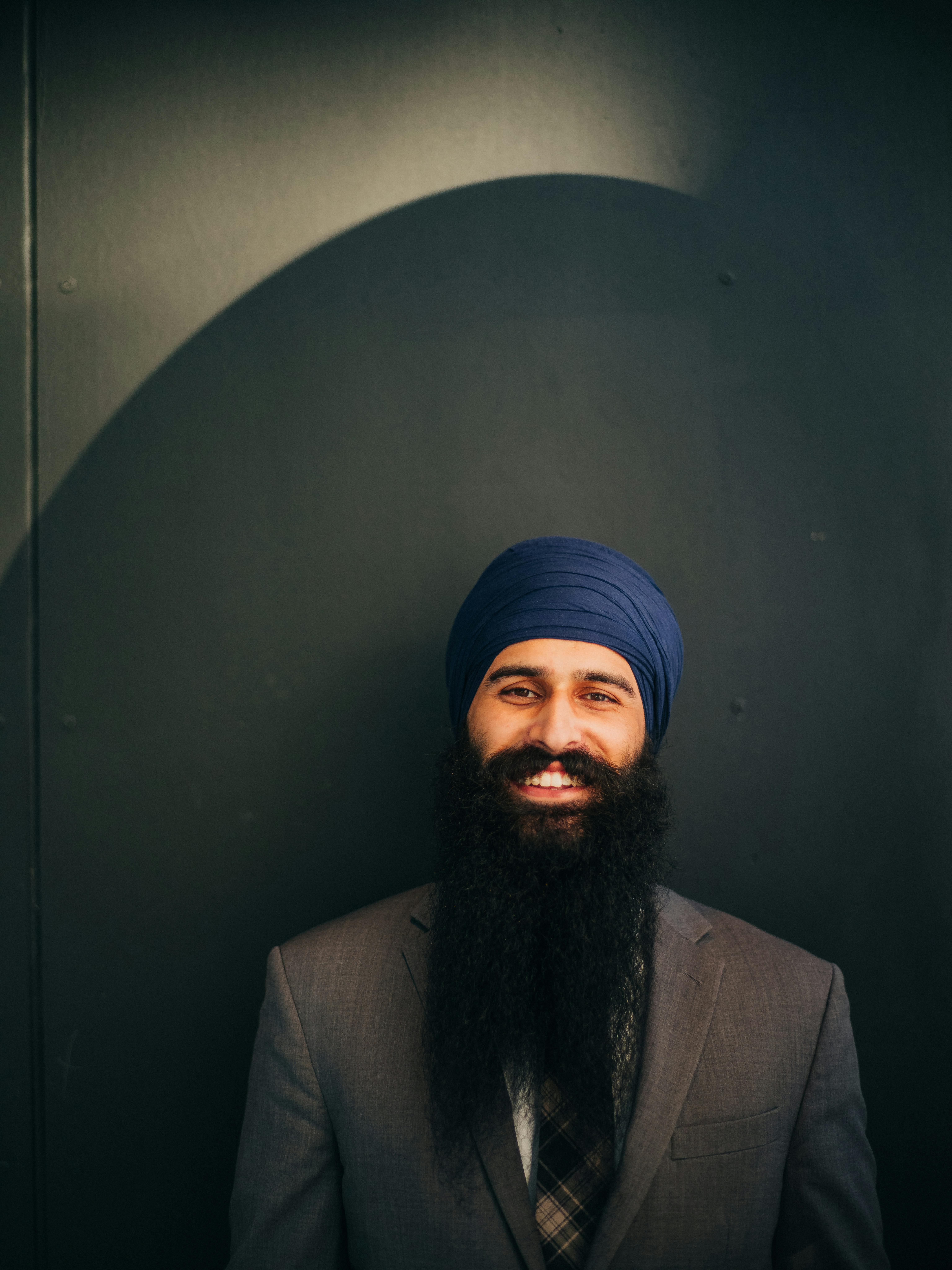 Bearded man wearing a turban smiling in front of a black wall.