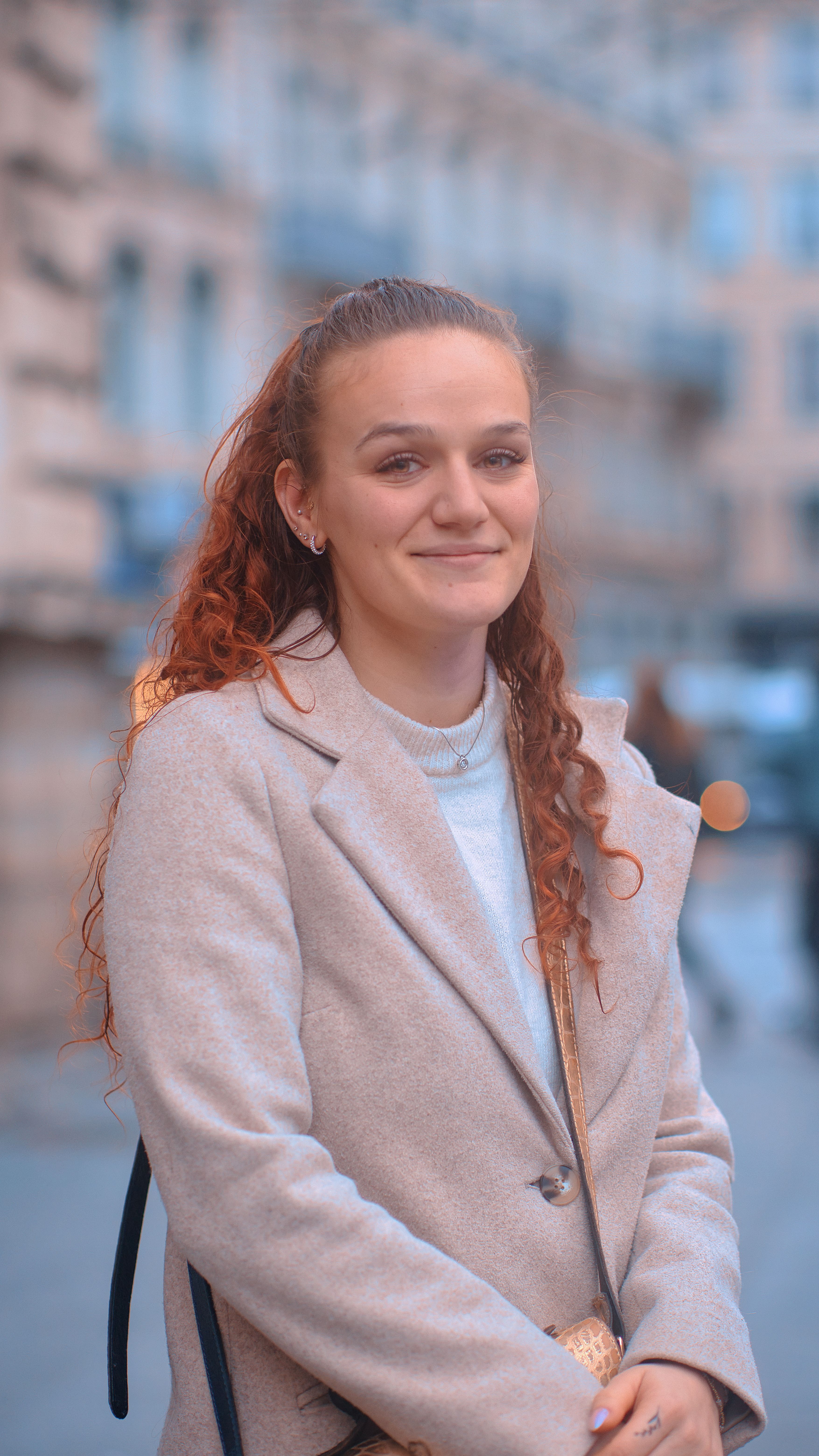 Portrait of a red-haired woman in a beige coat smiling confidently in an urban setting.