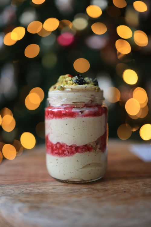 Free Delicious layered dessert in a jar with pistachio, raspberry, and cream against a bokeh light background. Stock Photo
