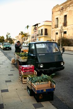 A street vendor with crates of fresh vegetables beside a vehicle on a busy urban street.