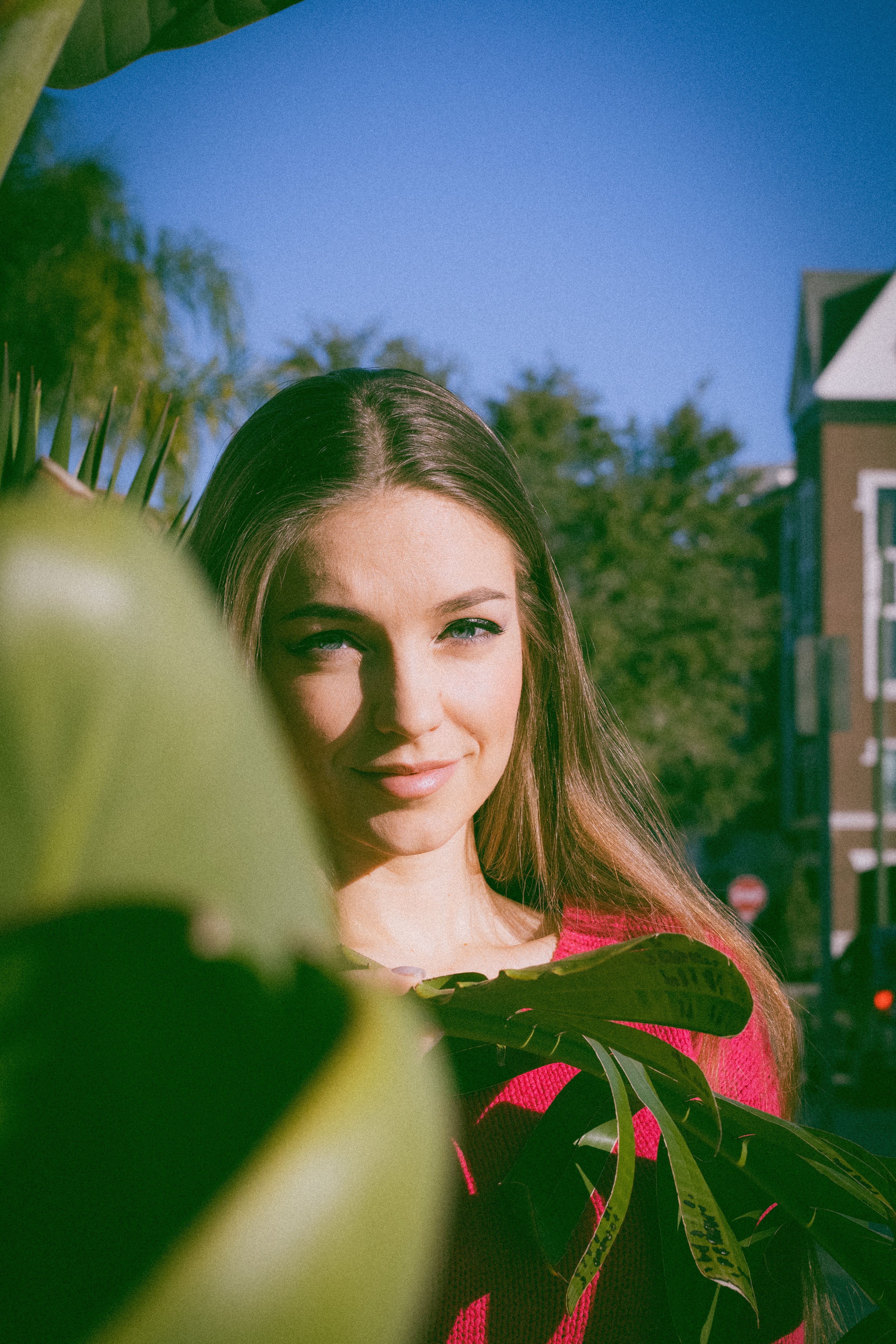 Smiling woman in red, partially obscured by leaves, on a sunny day.