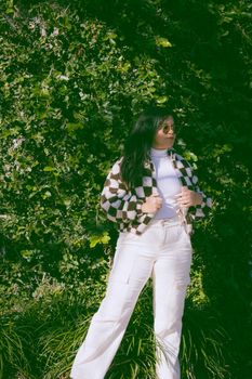 Fashionable woman in checkered jacket posing against vibrant green foliage outdoors in daylight.