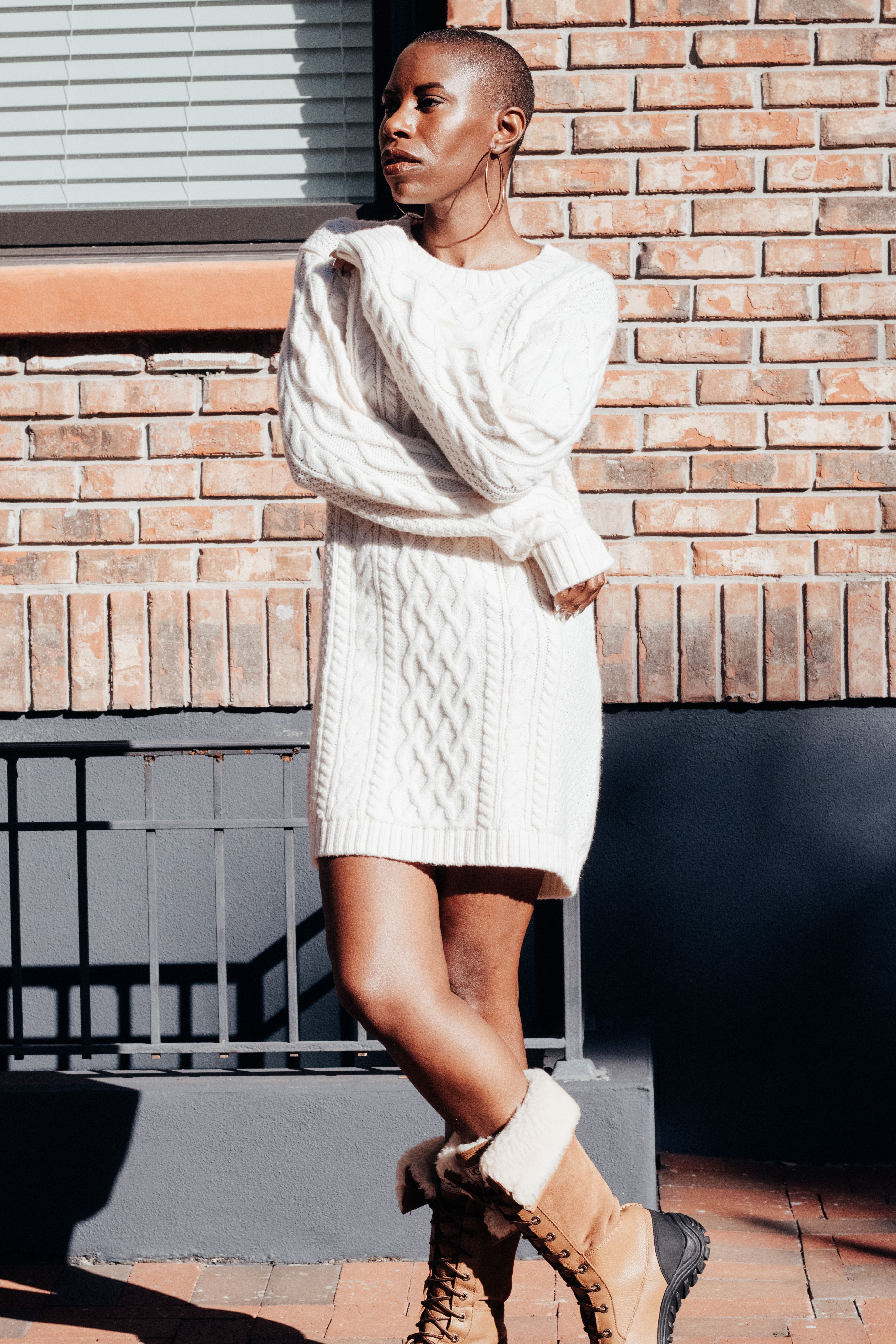 Woman pairing a cream cable-knit sweater dress with tan shearling-lined boots beside a brick wall
