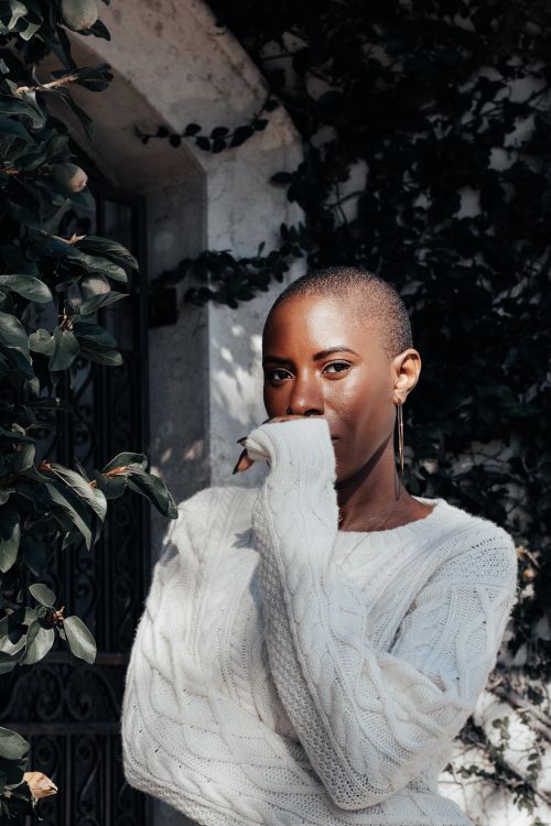 Free Stylish woman in a cozy white sweater stands confidently amidst lush greenery in bright sunlight. Stock Photo