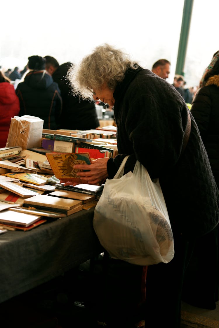 An Elderly Woman Looking At An Old Book On A Flea Market 