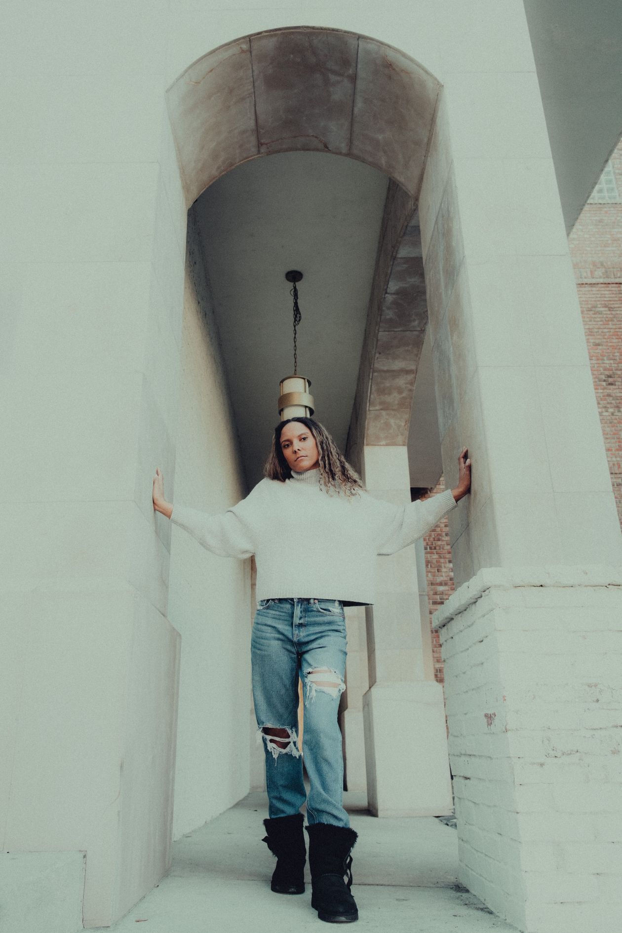 Woman in a white turtleneck and ripped jeans with black ankle boots standing under stone arches
