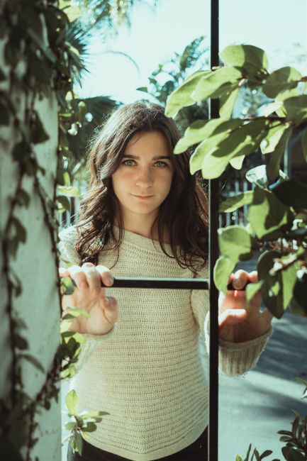 Young woman in a sweater posing outdoors with greenery, in natural light.