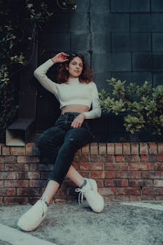Young woman in casual crop top and jeans, sitting against a brick wall outdoors, showcasing modern street fashion style.