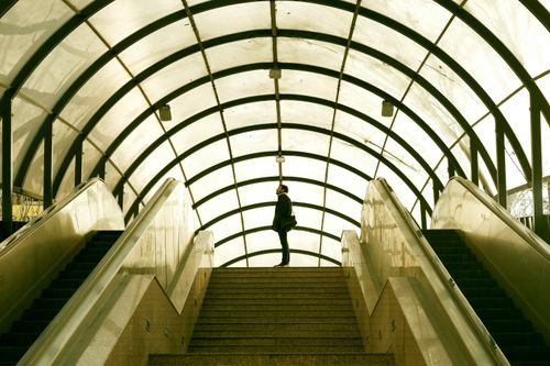Free A solitary figure stands in a glass tunnel above escalator stairs, creating a dynamic urban scene. Stock Photo