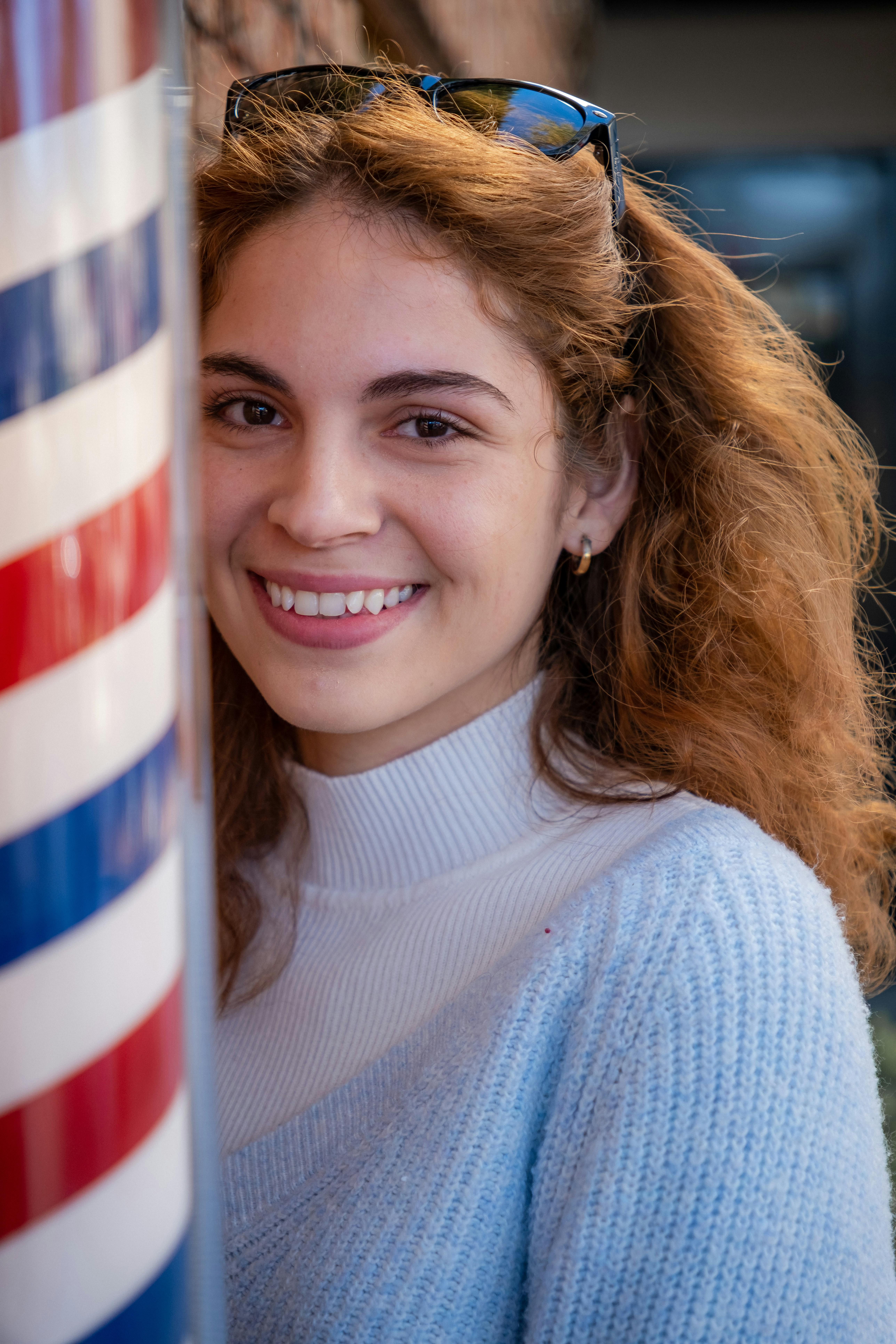 A young woman with curly hair smiling outdoors beside a barbershop pole.