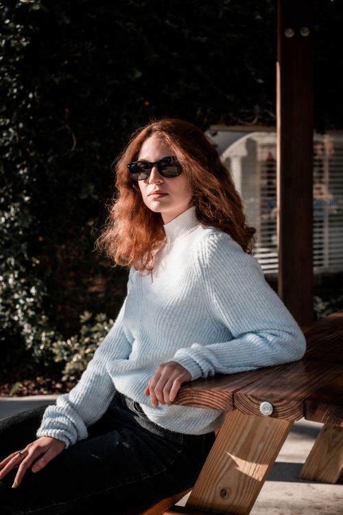Free Woman in white sweater and sunglasses, sitting on a bench, enjoying sunny outdoors. Stock Photo