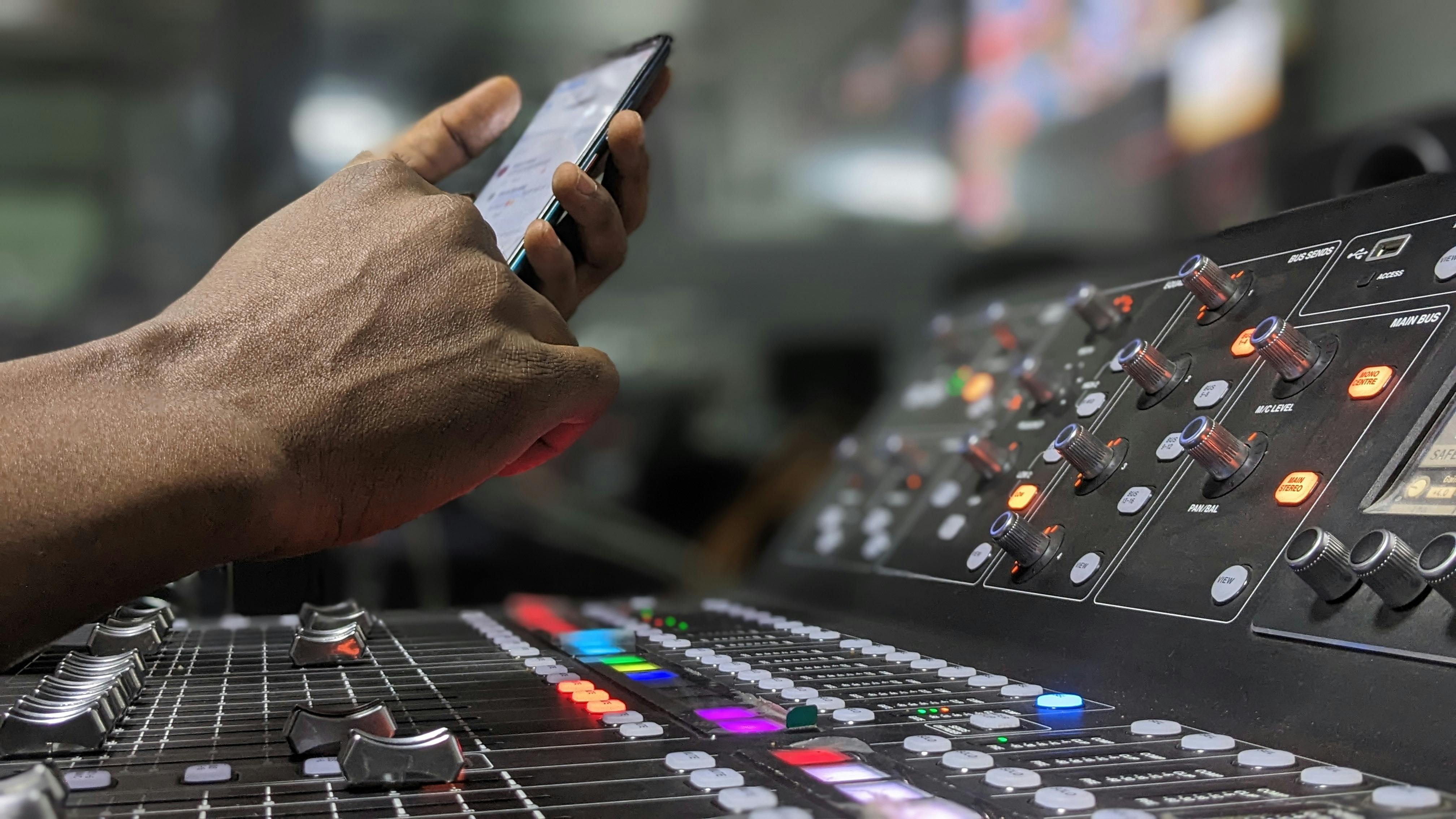 Close-up of audio engineer using smartphone and mixer controls in recording studio.
