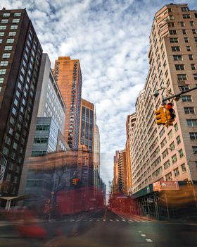 Dynamic city street scene with blurred motion of people and vehicles, framed by tall buildings under a cloudy sky.