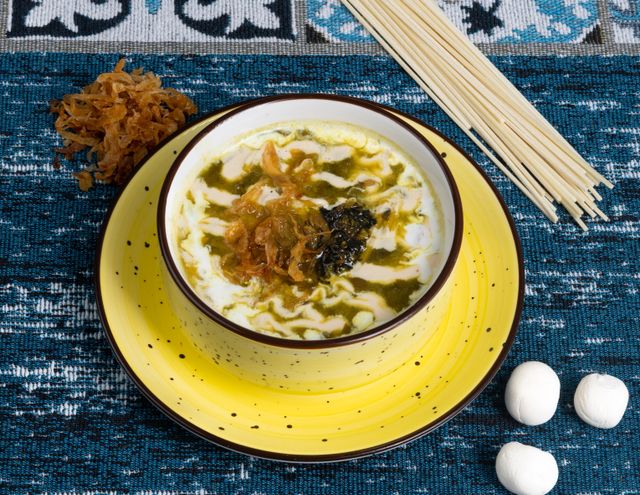 A vibrant soup with noodles and spices in a ceramic bowl on a decorative blue surface.