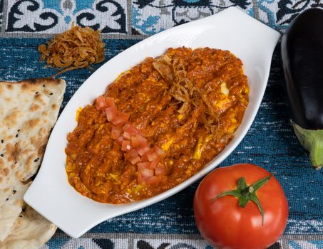 A vibrant dish of tomato and eggplant curry garnished with fried onions, served with bread.