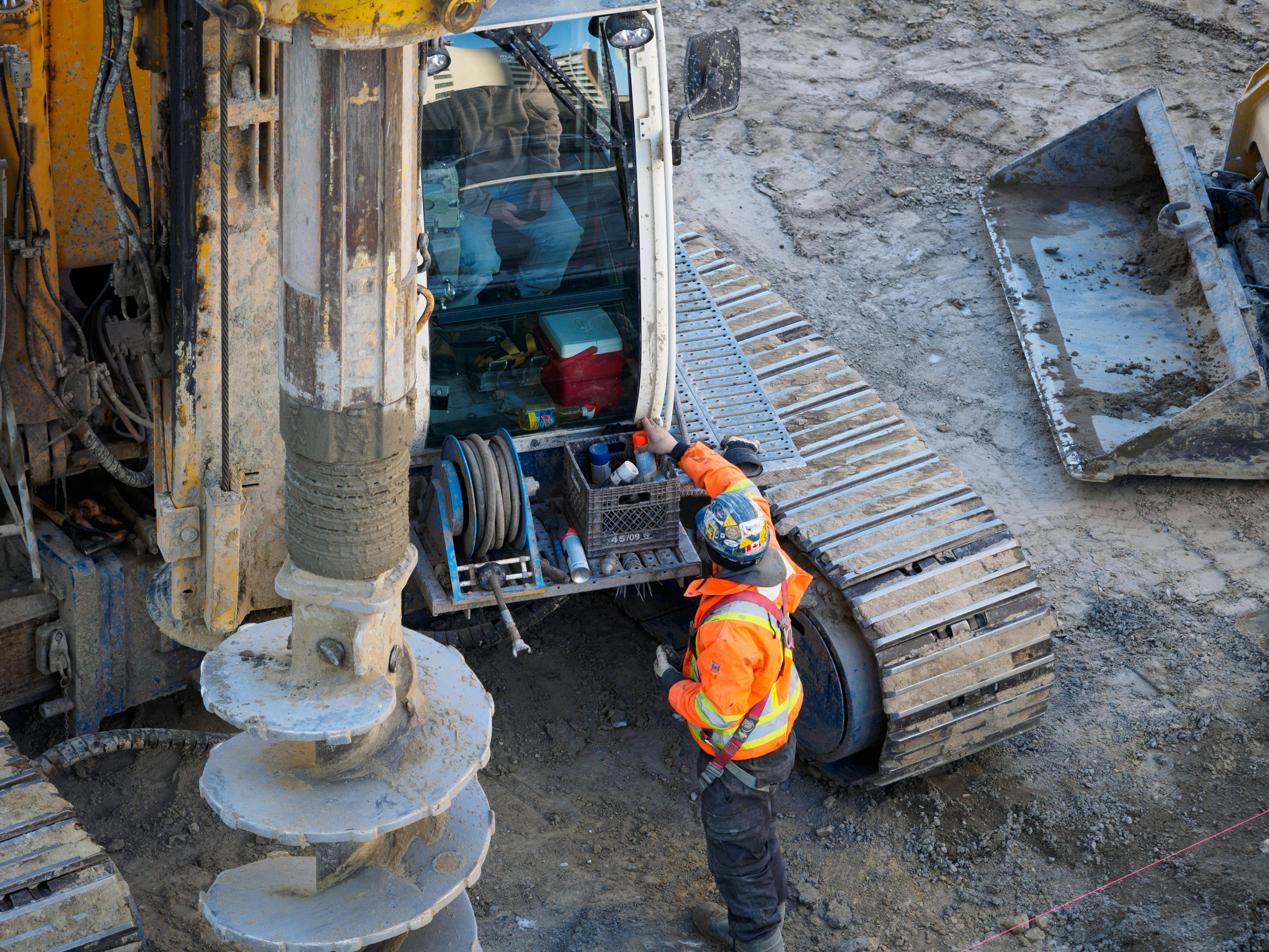 A construction worker in high-visibility gear operates heavy drilling equipment on a construction site.