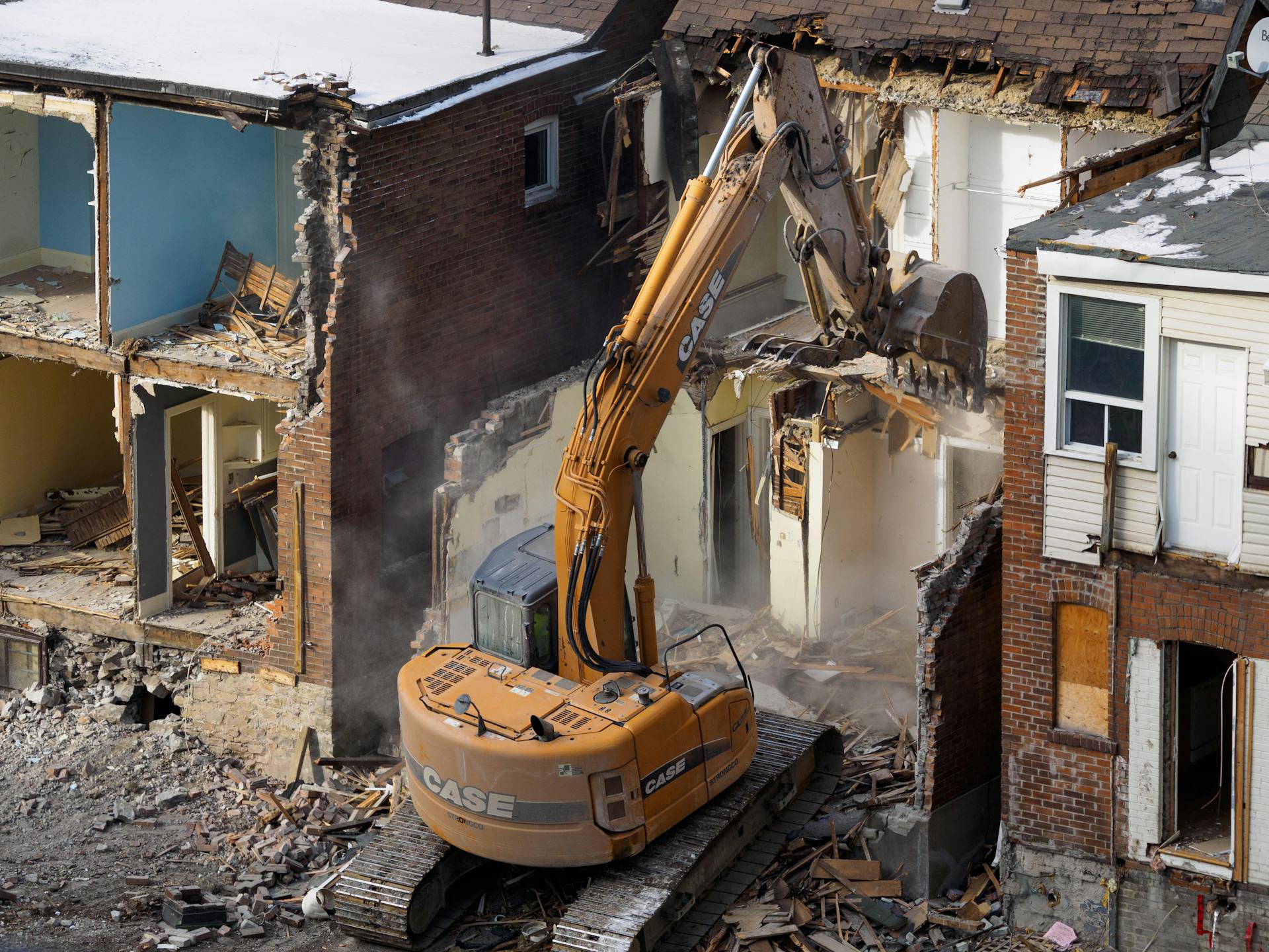 An excavator demolishes an old brick building at a construction site, creating debris and dust.