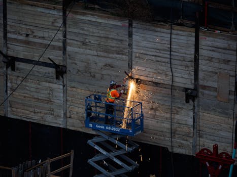 A construction worker welds on a raised platform, creating sparks on an industrial site.