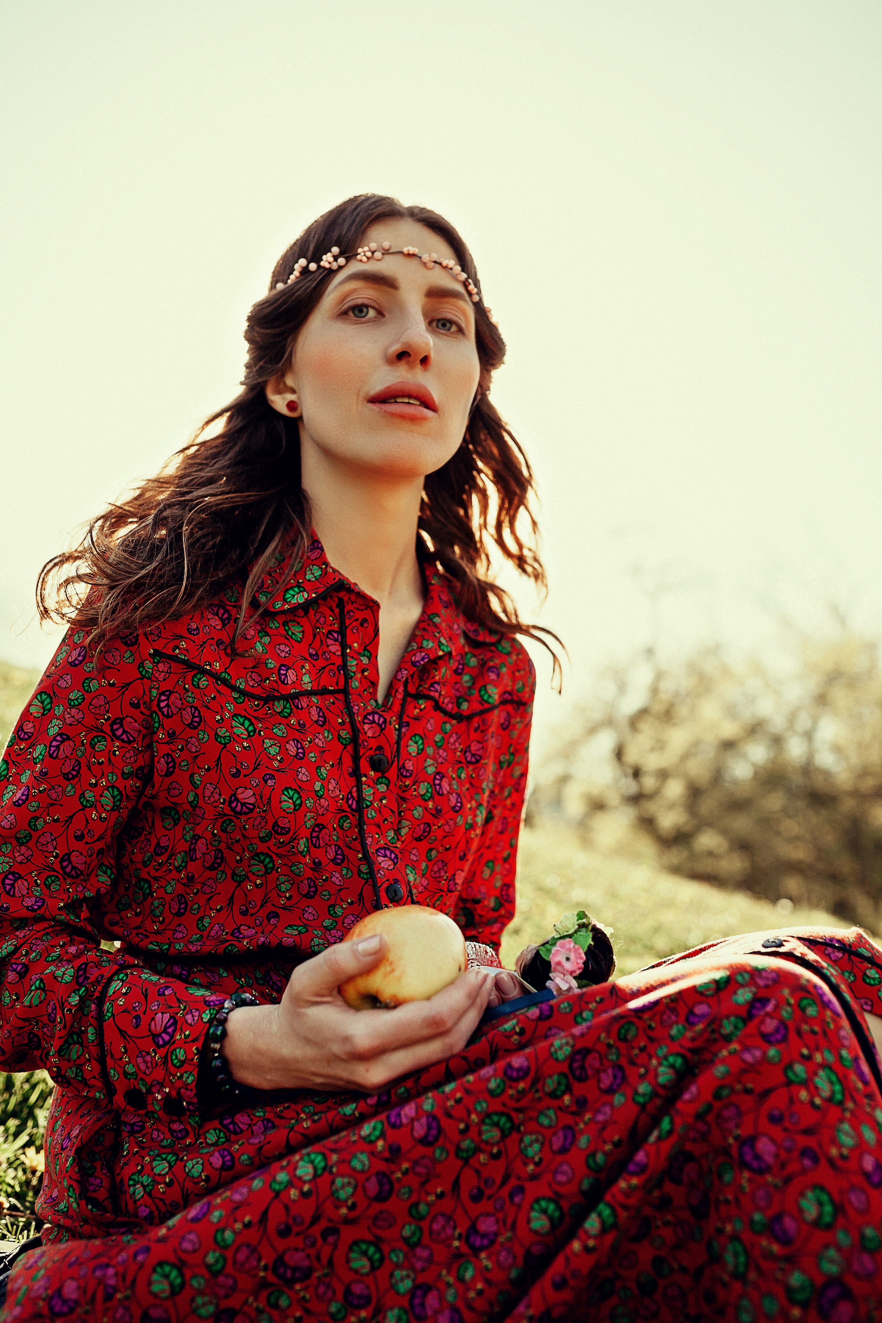 A woman in a vibrant floral dress sits outdoors holding an apple, enjoying the summer sun.