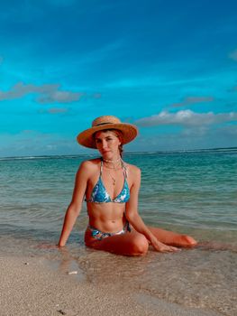 Woman in a bikini and straw hat sitting on a sandy beach by clear ocean water under a blue sky.