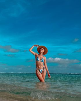 A woman in a bikini and hat confidently poses at a tropical beach, showcasing summer fashion.