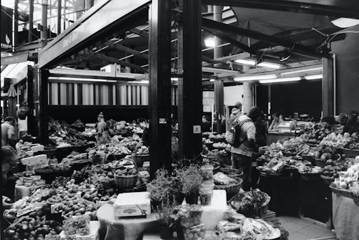 A bustling black and white photo of a busy market in England showcasing a variety of fruits and vegetables.