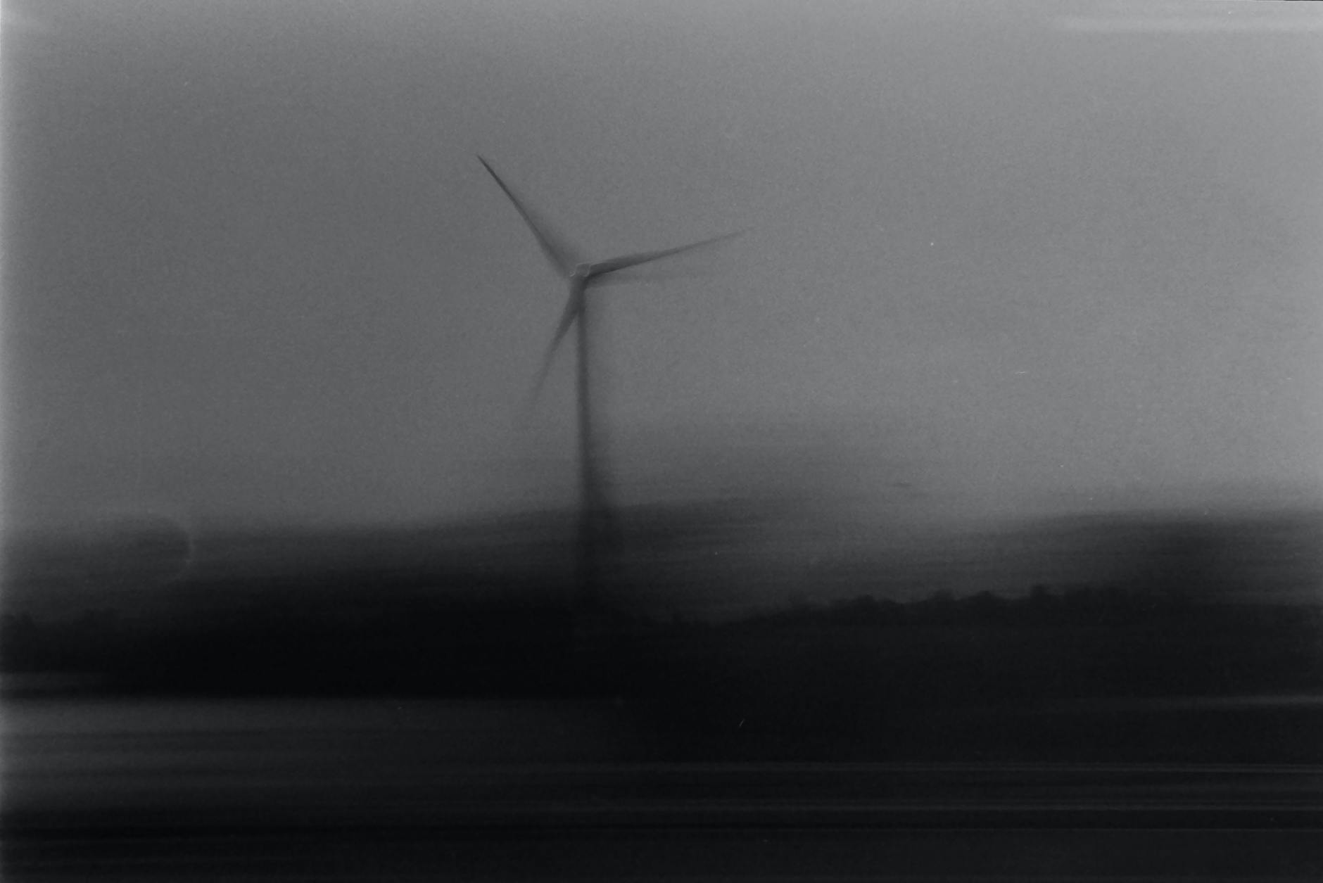 Blurred black and white image capturing the motion of a solitary wind turbine, creating an eerie feel.