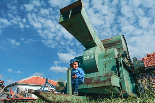 Free A young child sits on rustic farm machinery in a rural setting under a blue sky with scattered clouds. Stock Photo